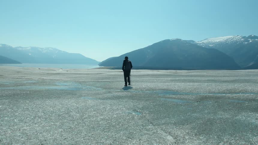 A lone hiker walks across a vast icy landscape with mountains in the background