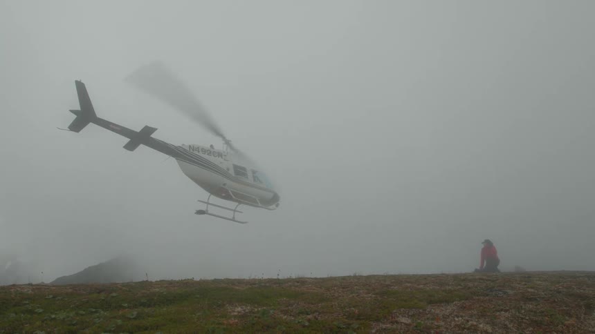 A helicopter is flying and went away through fog while a person wearing red jacket weaving his hand