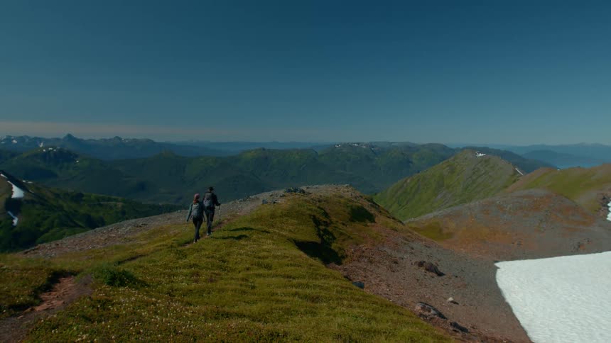 Two hikers walk along a grassy ridge overlooking a vast mountain range with lingering snow patches in the foreground
