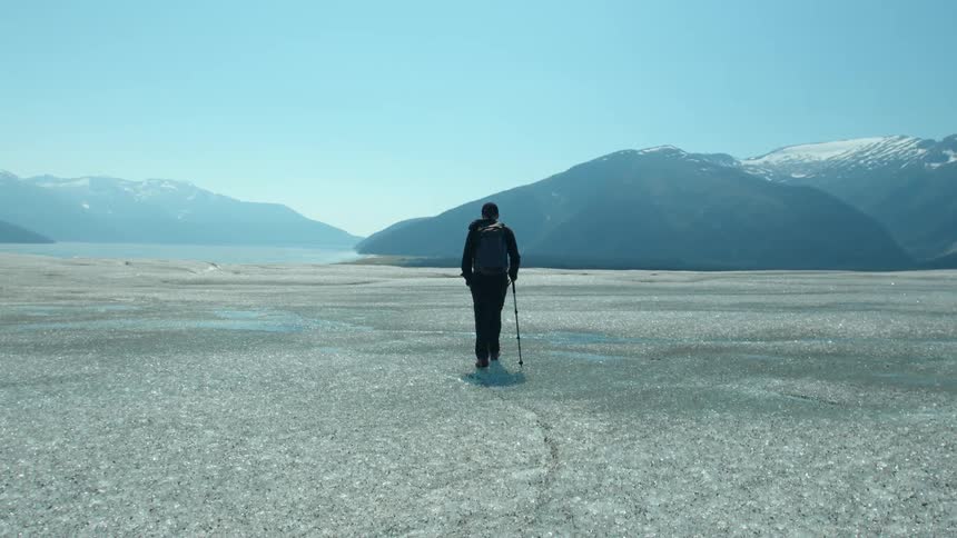 A lone hiker walks across a vast icy landscape toward distant snow covered mountains