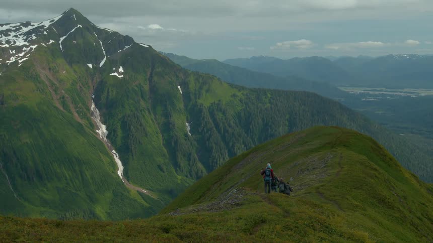 Hikers walk on a green ridge with snowy peaks and waterfalls behind