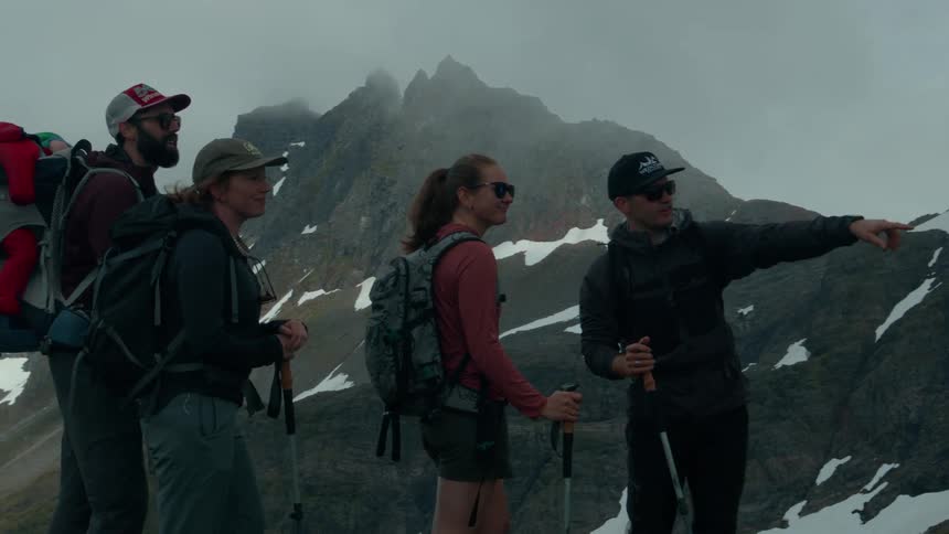 A group of hikers with backpacks pauses on a rugged snow patched mountainside as one person points towards a dramatic mist shrouded peak