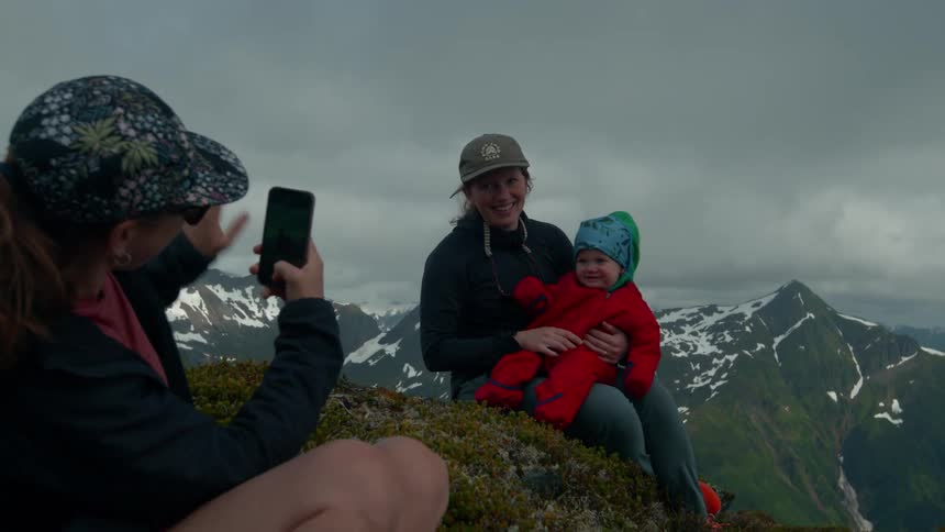 Woman holds a baby while another woman takes a photo on a mountain summit