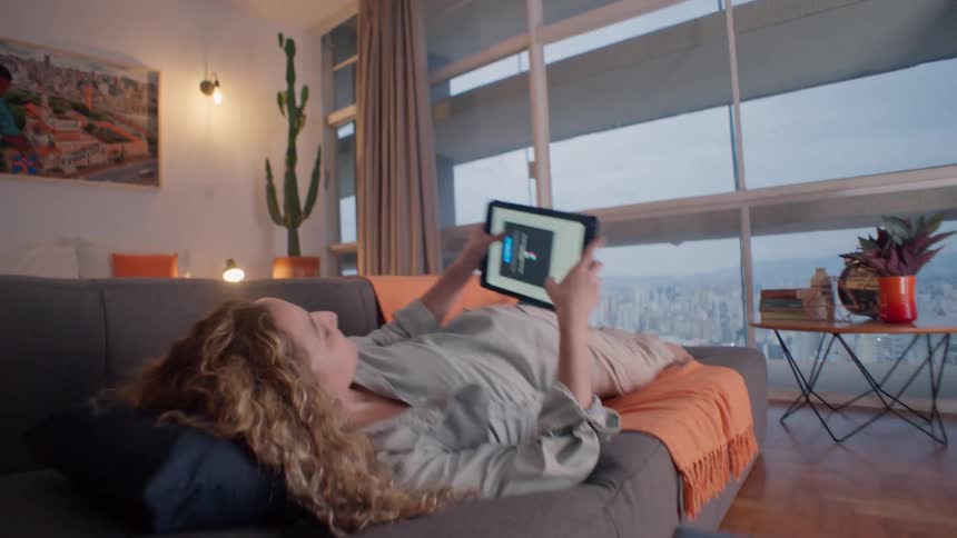 Woman lounges on sofa using tablet by cityview windows