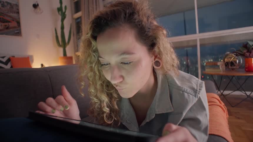 Close up of a young woman smiling and scrolling on a tablet computer while laying on a couch in a living room