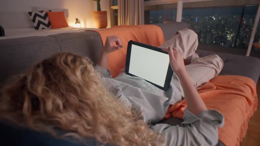 A young woman uses a tablet computer while lying down on a couch in a living room at night