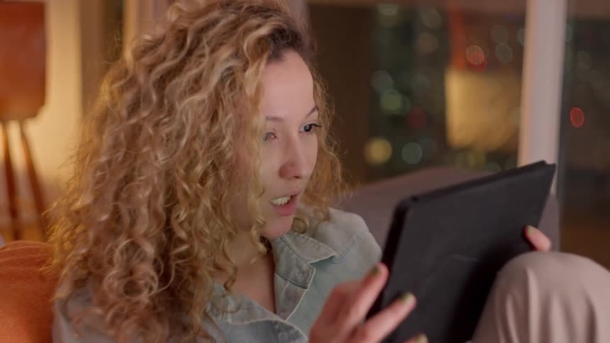 A woman relaxes on a couch while using a tablet illuminated by warm indoor lighting