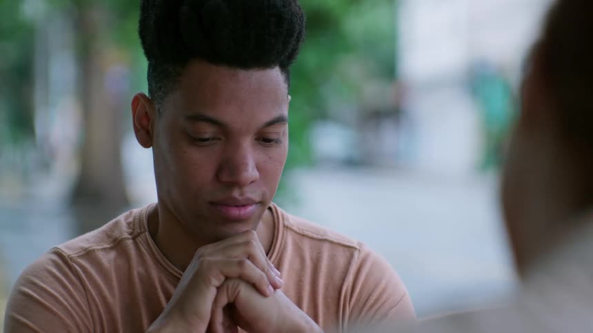 A young man sits thoughtfully with his hands clasped under his chin attentively listening to someone across from him in an outdoor setting