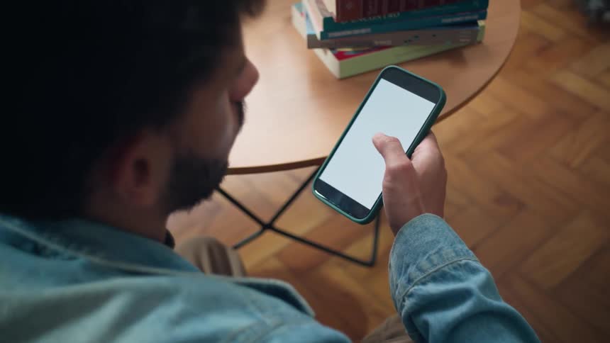 A young man sits in front of a coffee table scrolling on his smartphone with a few books stacked on the table
