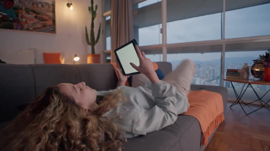 A young woman lies on a couch in a living room to use a tablet computer