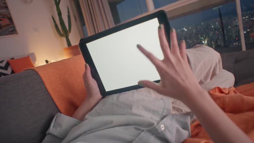 Person relaxing on a couch at night while using a tablet with a blank screen