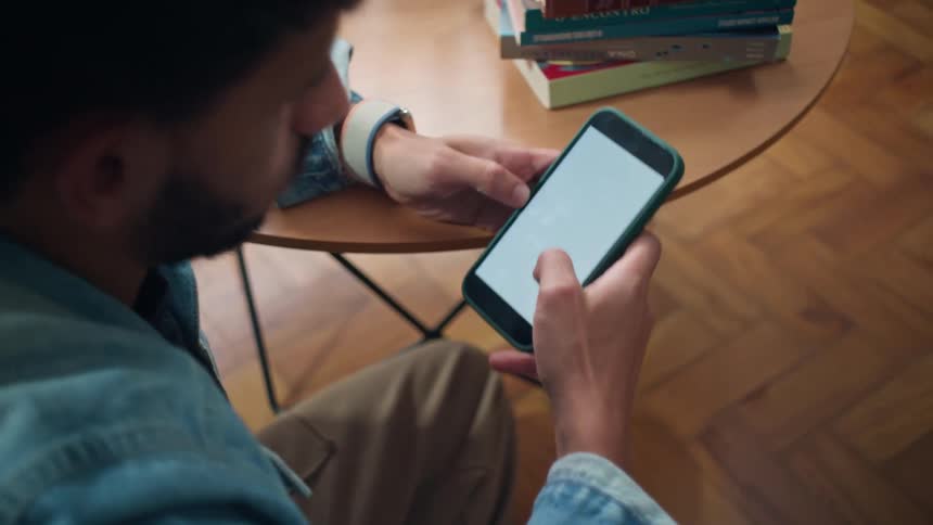 A man sits at a table intently looking at a smartphone with a blank white screen while resting his hand near his mouth