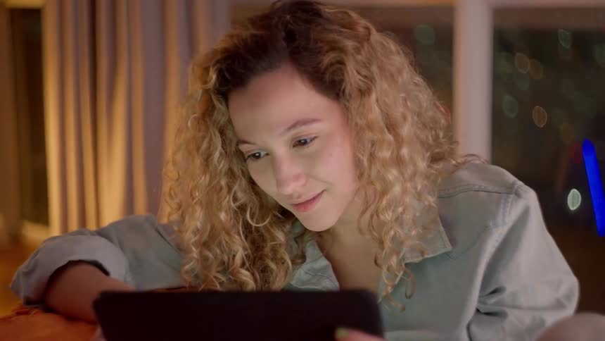 A woman with curly hair smiles while looking at a tablet screen indoors at night