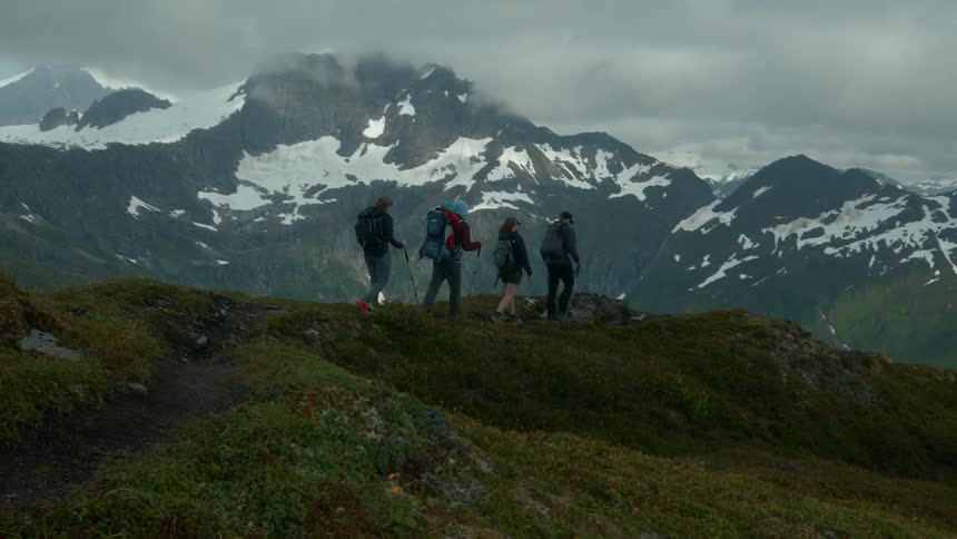 Hikers trek across an alpine ridge with snowy mountains in the background