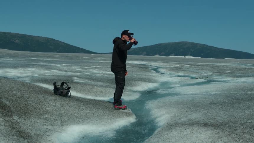 Tourist kneels to fill bottle from a glacial stream on icy terrain
