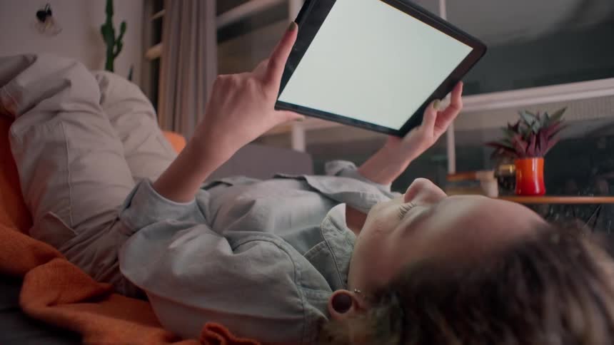 A woman lies on a couch holding a tablet with a blank screen
