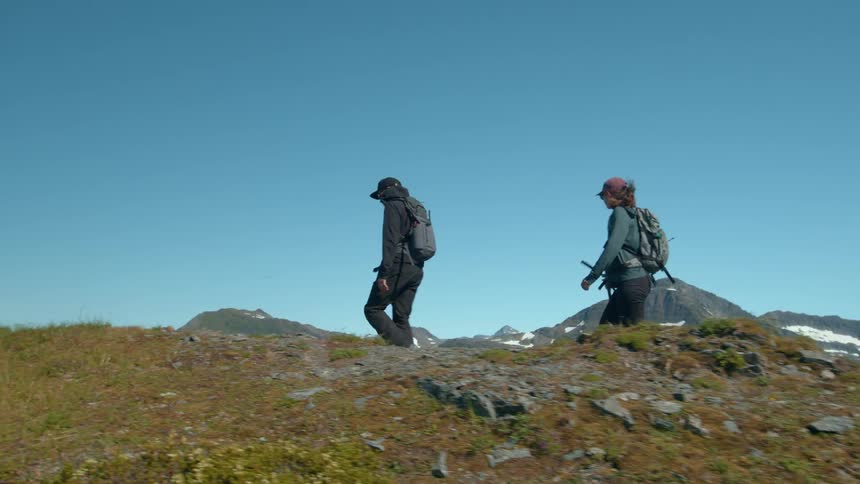 Two hikers trek across a scenic mountain trail under a clear blue sky