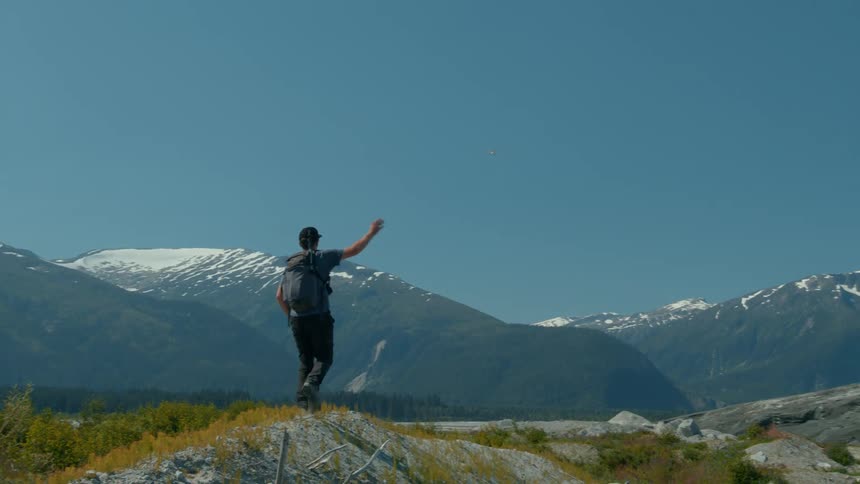 A male hiker with a backpack standing on a ridge with snowcapped mountains in the background he then raises his right hand in a wave or salute towards the distant scenery