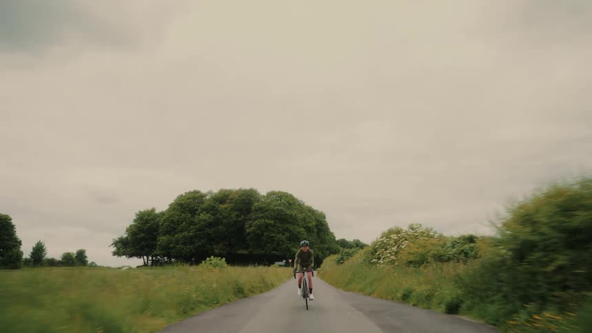 A cyclist rides down a quiet country road flanked by green fields and trees