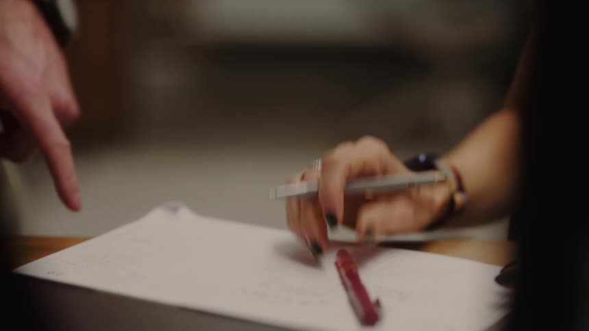 Using a pen a woman writes a note on a paper while a male companion points his finger