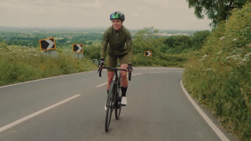 A cyclist rides uphill on a winding country road surrounded by greenery