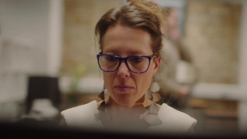 A woman in blueframed glasses and a white top works at a computer in an office while two men converse in the blurred background