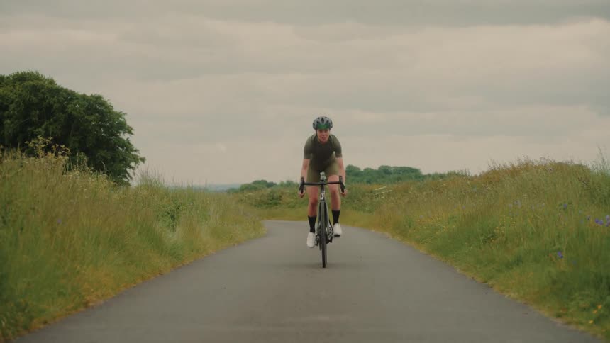 Female cyclist smiles while riding a bicycle on a paved road passing overgrown fields under an overcast sky on a summer day