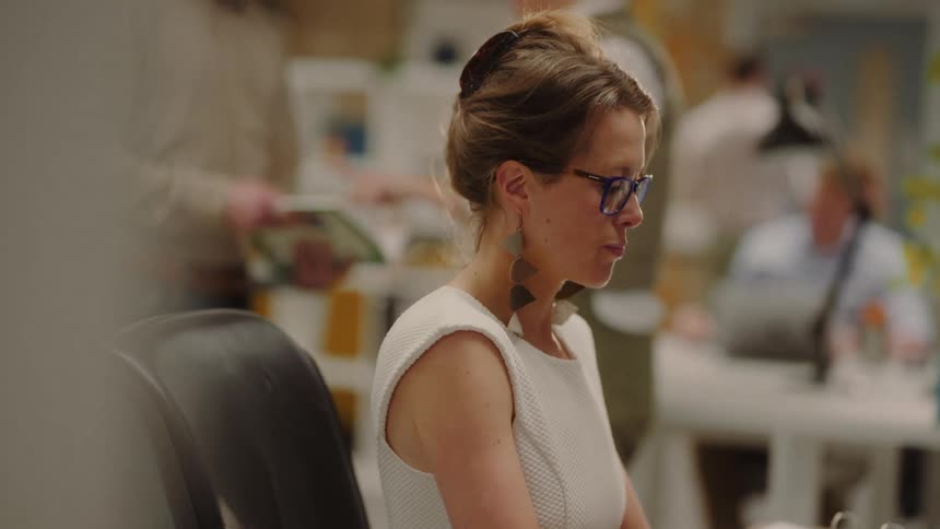 A woman with hair pulled back wearing a white sleeveless top and blue framed glasses works in a busy office space