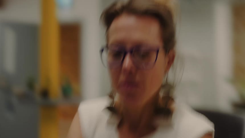 A person wearing glasses is seen in an office setting initially typing on a laptop with a blurred background the camera then focuses on their face as they answer a phone call shifting from work to communication
