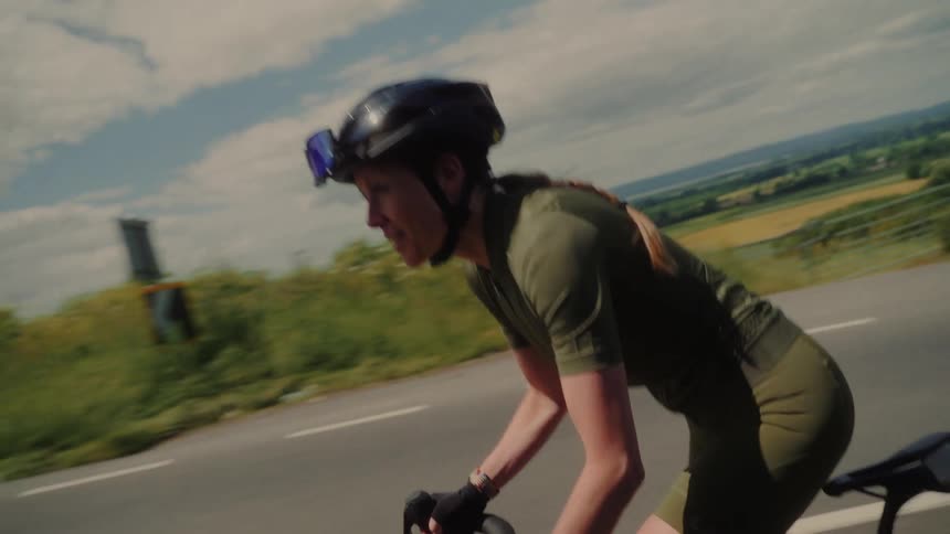 A female cyclist wearing a helmet and green cycling gear riding a black road bike on a scenic road she is actively pedaling with a focused expression against a backdrop of rolling hills under a cloudy sky
