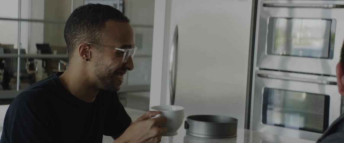 A man sits in a kitchen laughing and drinking from a mug