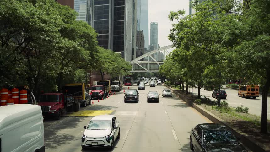 Cars drive through a city street lined with trees and tall buildings