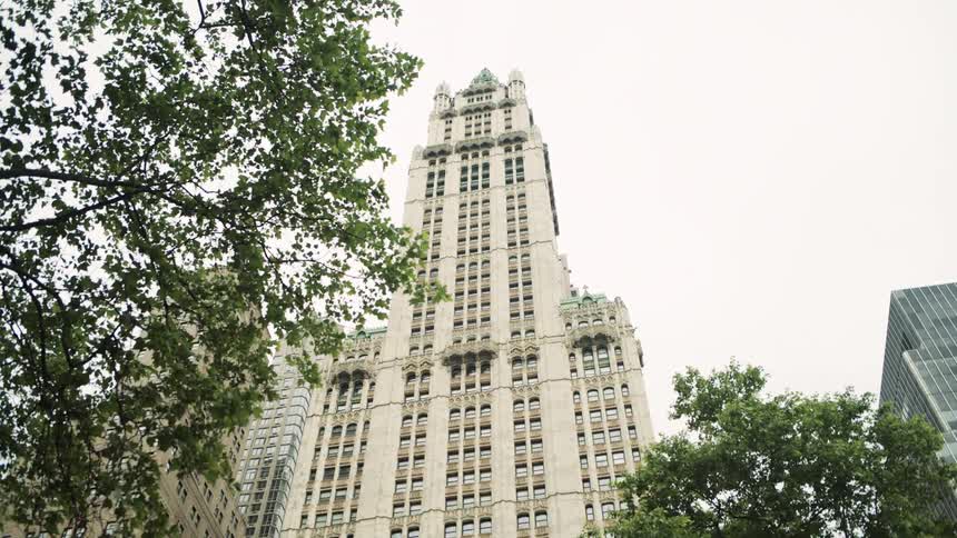 A lowangle shot looks up at an ornate historic skyscraper in a dense city with its facade framed by the green leaves of nearby trees