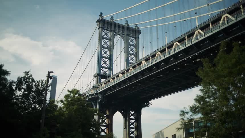 Under a pale sky the manhattan bridge stretches across the frame its dark metal framework and crisscrossing cables visible above city buildings and trees below