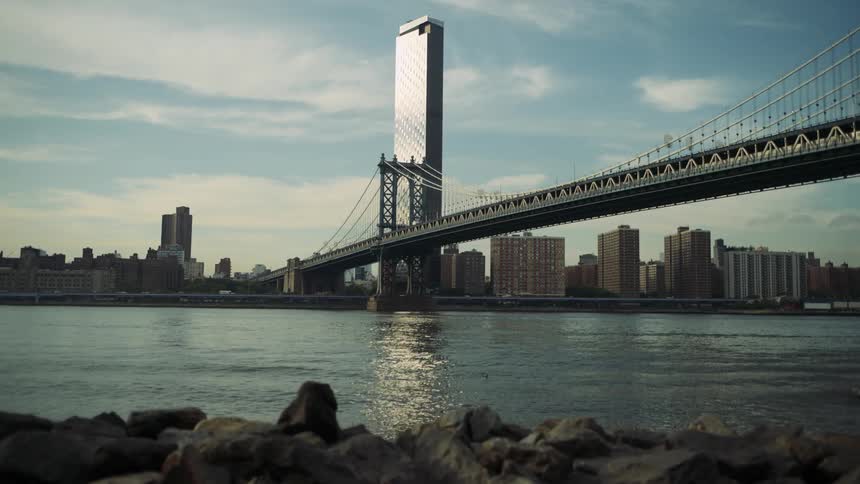 A suspension bridge stretches over a calm river with a city skyline in the background
