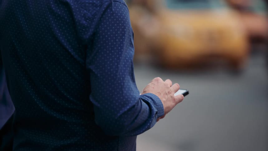 A man stands on the sidewalk holding and operating a mobile device as people walk by and cars pass on the street at midday