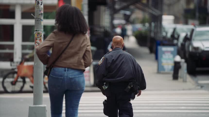 A bald man in a police uniform walks through a city crosswalk surrounded by pedestrians