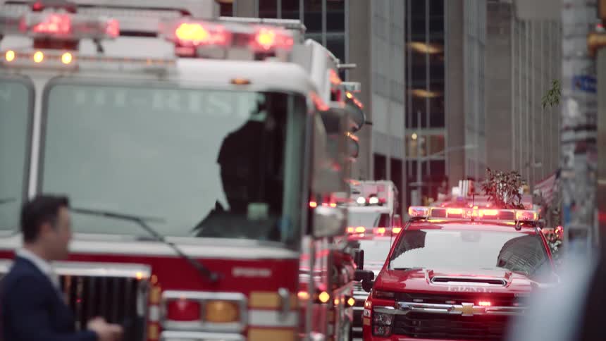 A line of red fire trucks and a red pickup truck all with flashing lights are parked on a city street with buildings in the background