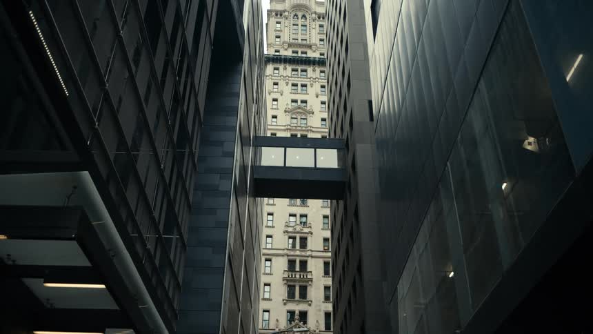 A lowangle view looking up a narrow urban canyon formed by two modern darkcolored buildings on either side in the distance framed between these two structures a classicalstyle lightercolored building with numerous windows rises high into the sky