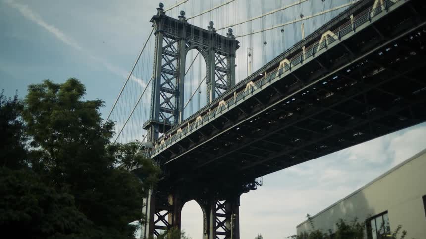 A lowangle shot from below captures the immense steel structure of the manhattan bridge against a partly cloudy sky with trees framing the view