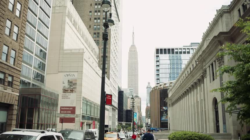 A street view of a city with tall buildings lining both sides of the road in the background a prominent skyscraper likely the empire state building can be seen towering over the other structures
