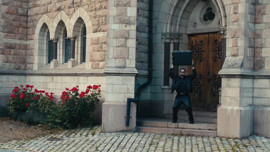 Man stands outside a stone building holding a suitcase overhead while acting funny