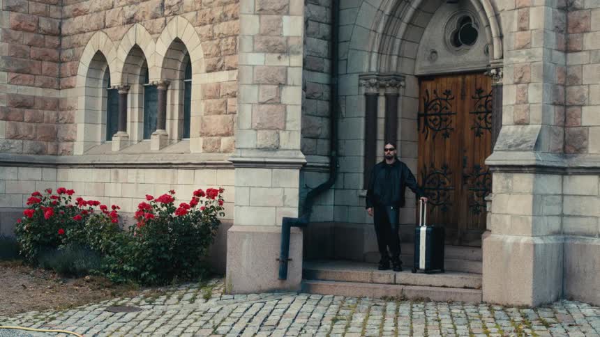 A man with a suitcase stands outside a stone building near a wooden door