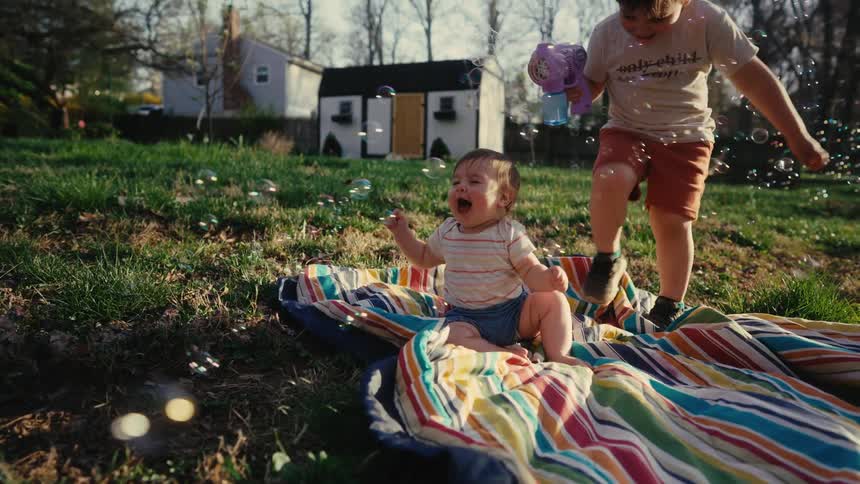 Baby sitting on a colorful rug on the grass with soap bubbles flying and another child next to it