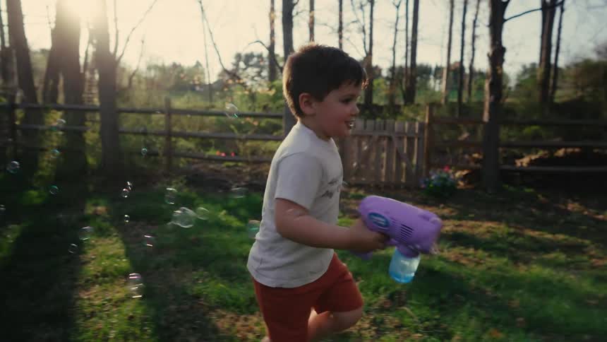 A young boy runs through a sunlit backyard holding a bubble gun