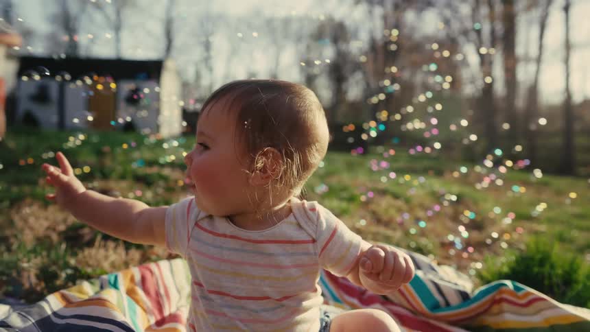 A baby sits on a blanket outdoors watching bubbles float by in a sunny backyard