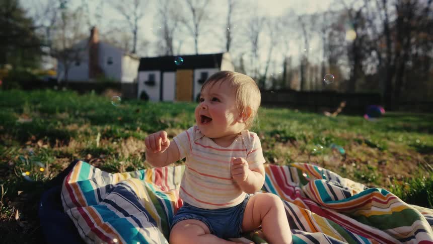 A baby sits on a blanket outside and watches in awe as bubbles float by him
