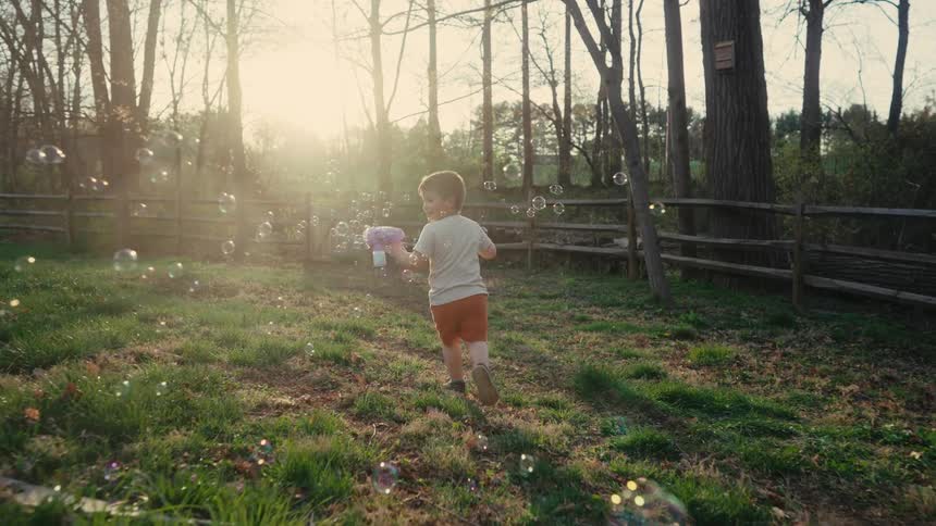 Child runs across the lawn blowing soap bubbles as the sun sets behind the trees