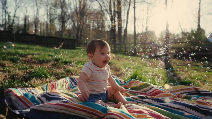 Little girl sitting on a colorful towel with a happy expression and a boy blowing soap bubbles