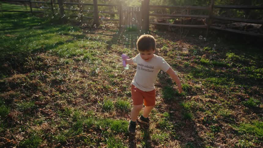 A young boy plays with bubbles in a sunlit yard surrounded by grass and enclosed by a rustic wooden fence